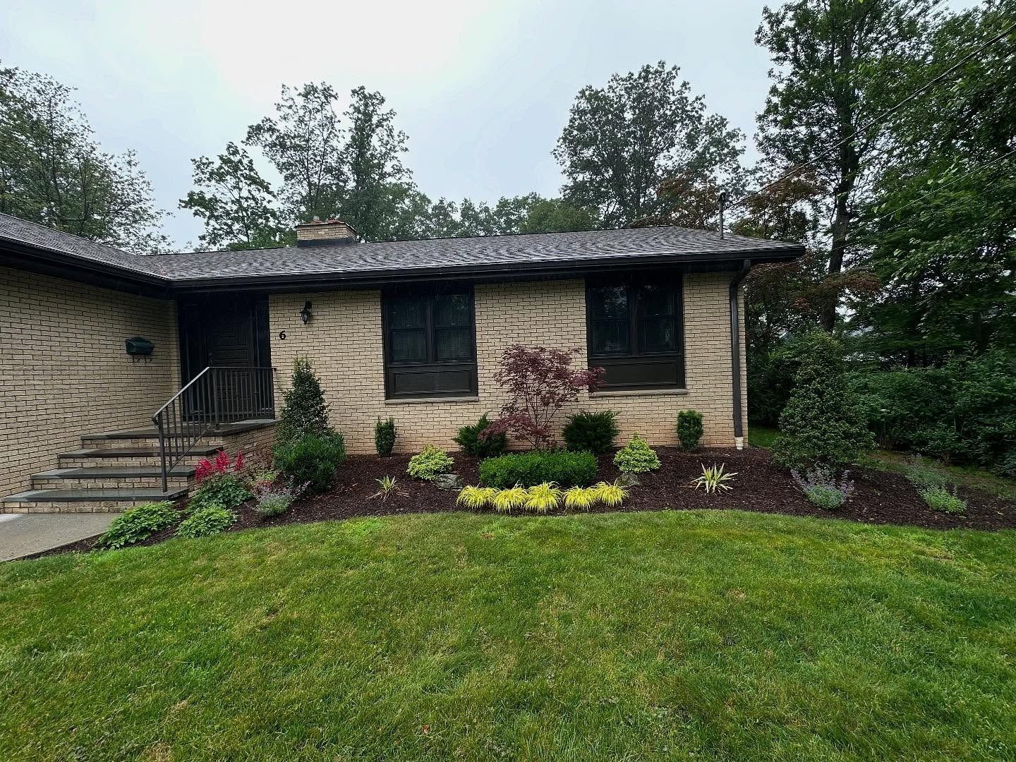 Single-story stone house with a dark roof, front steps, and a manicured green lawn with shrubs and flowers.