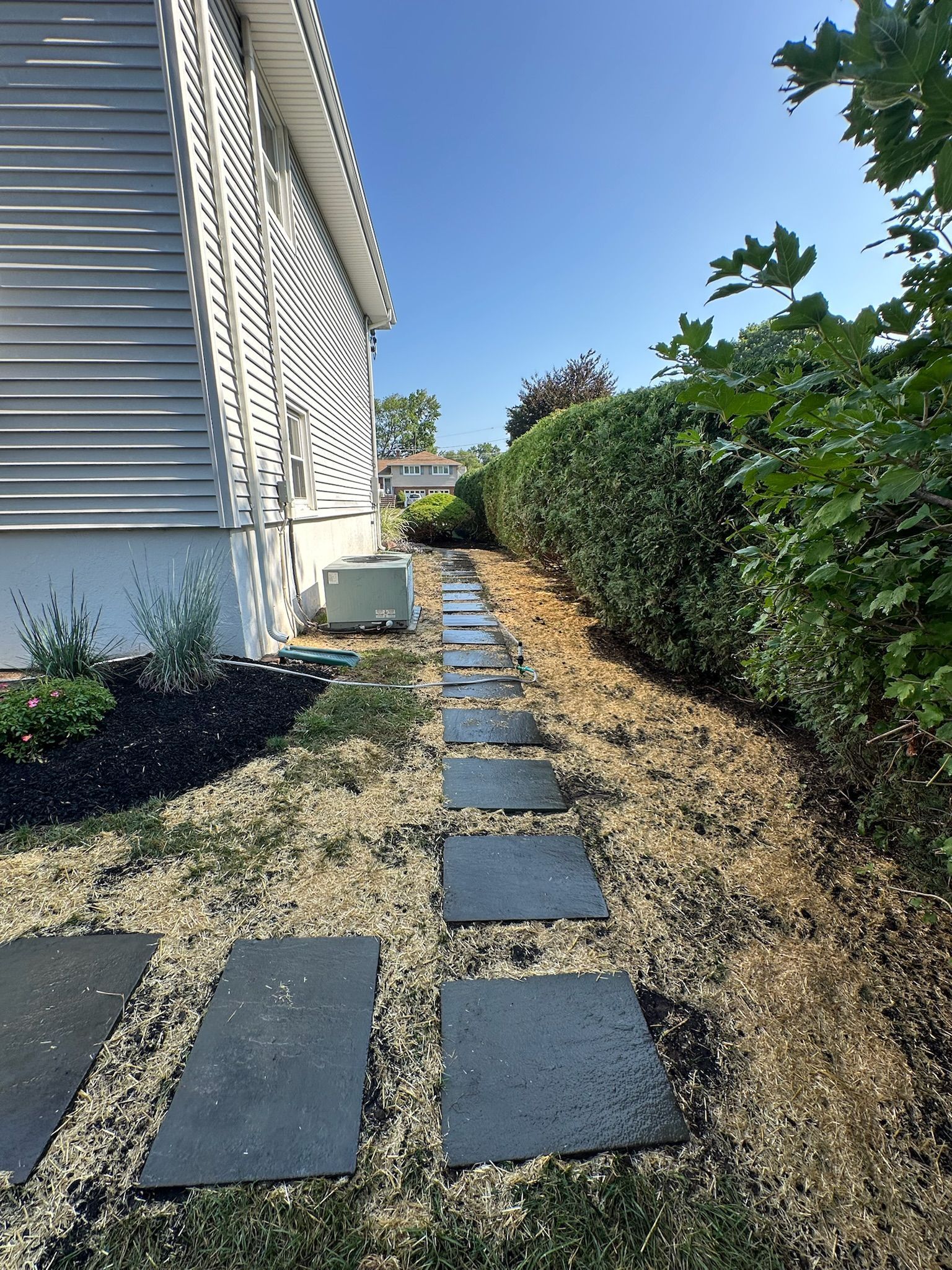 Stepping stones along a narrow side yard beside a house, with hedges and gravel under a blue sky