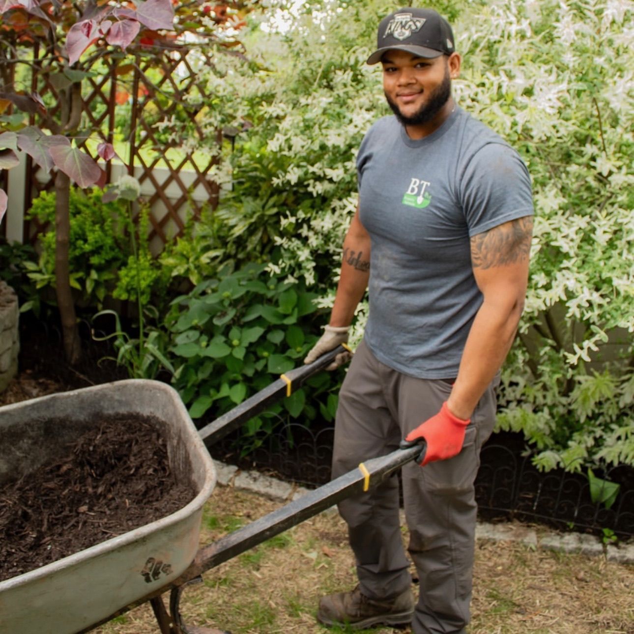 Gardener in gray shirt and gloves pushes a wheelbarrow in a lush backyard garden.