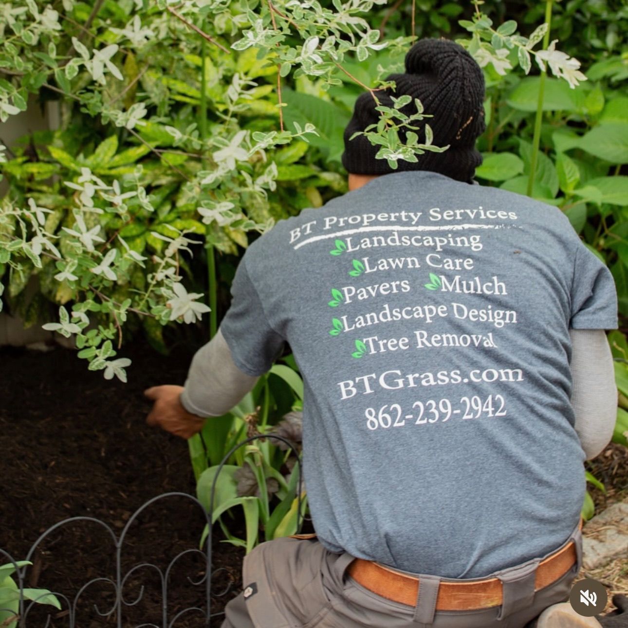 Landscaper in gray shirt kneels by bushes, pruning greenery in a garden.