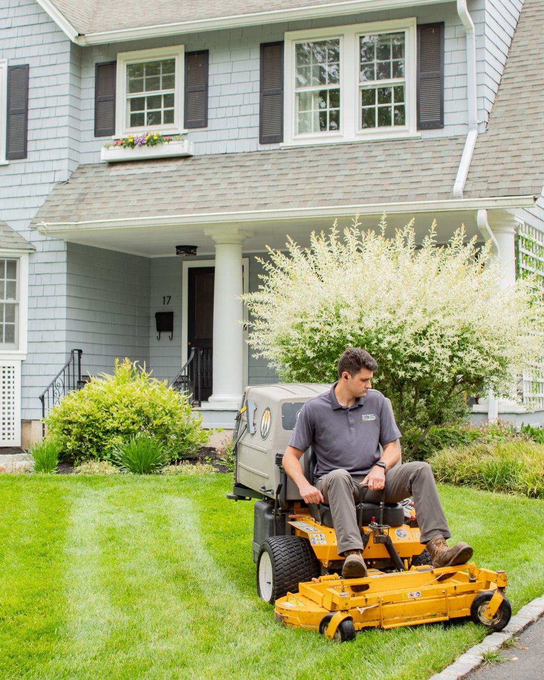 Man mowing a bright green lawn with a yellow riding mower in front of a house