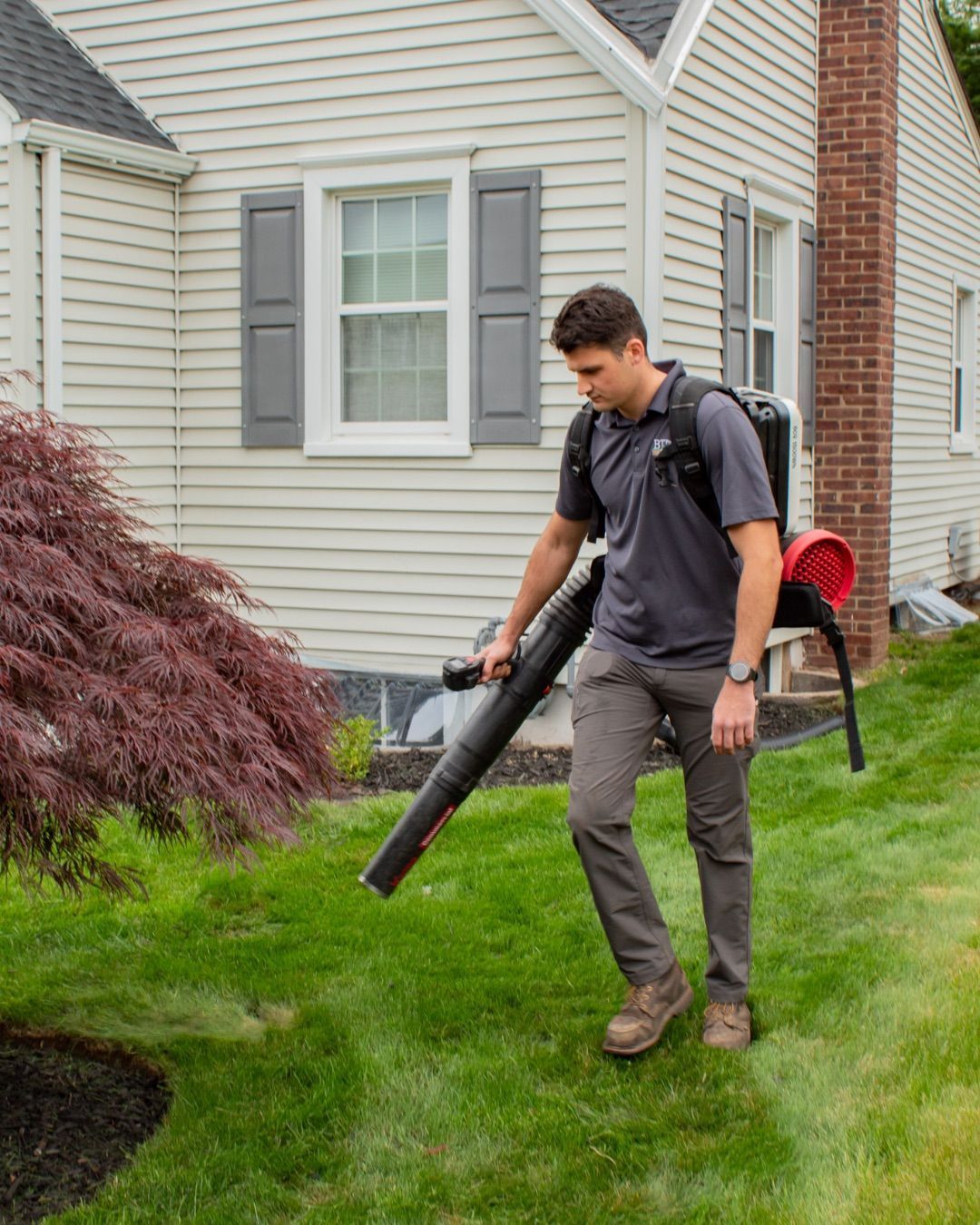Person using a leaf blower on a lawn beside a beige house with gray shutters