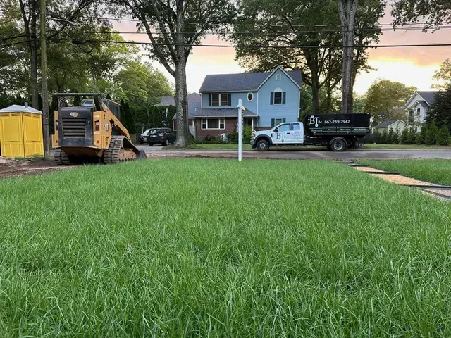 Lawn in foreground with dump trucks and a blue house on a tree-lined street at dusk