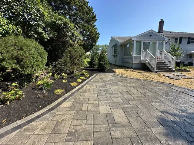 Paver driveway beside a small white house with a front porch, landscaped beds, and trees on a sunny day