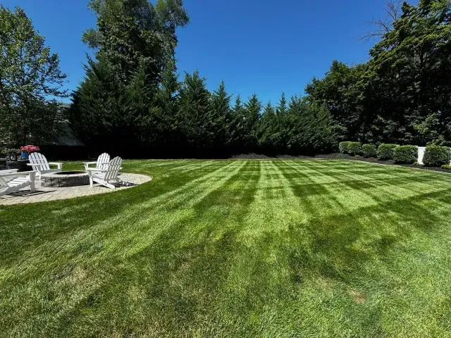 Wide striped green lawn with white patio chairs beside a hedge under a bright blue sky.