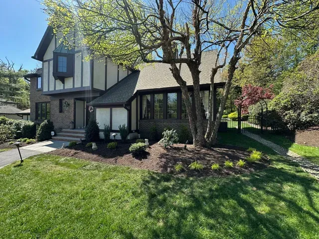Suburban two-story house with Tudor-style accents, trees in front yard, and green lawn in sunlight