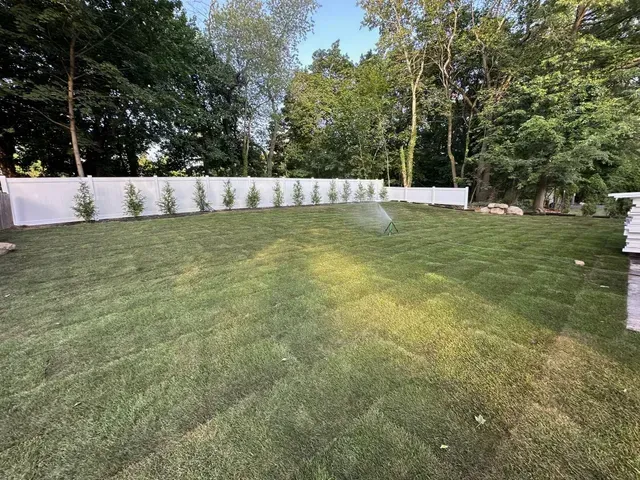 Backyard lawn with a white fence, small shrubs, and trees in the background