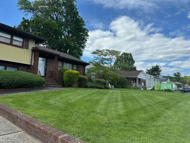 Suburban homes with a wide green lawn, trees, and a partly cloudy blue sky