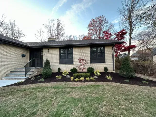Single-story beige house with dark roof, front lawn, and landscaped shrubs under a clear sky