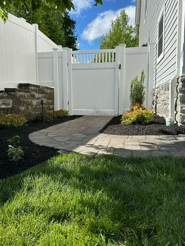 Stone garden path leading to a white gate and fence beside a house, with grass and flowers in sunlight
