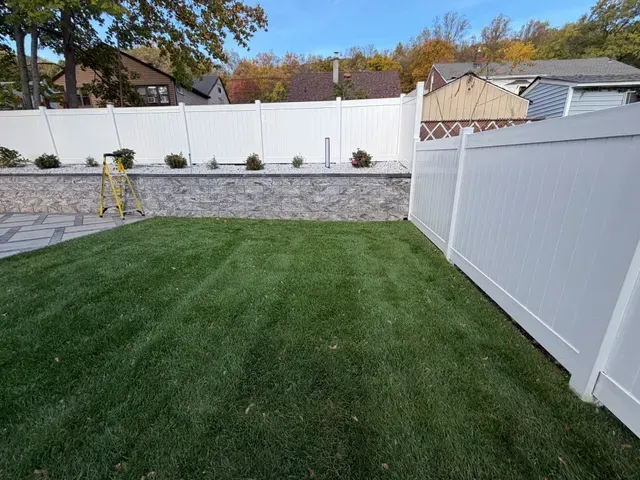 Backyard with green lawn, white vinyl fence, stone retaining wall, and small patio area