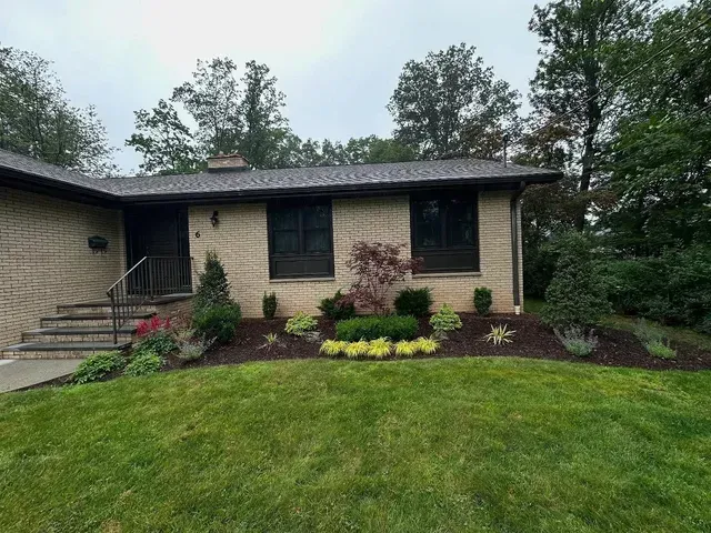 Single-story brick house with front porch, dark roof, and landscaped flower beds on a green lawn.