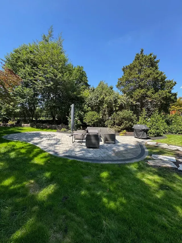 Backyard patio with fire pit, chairs, and lush green trees under a clear blue sky