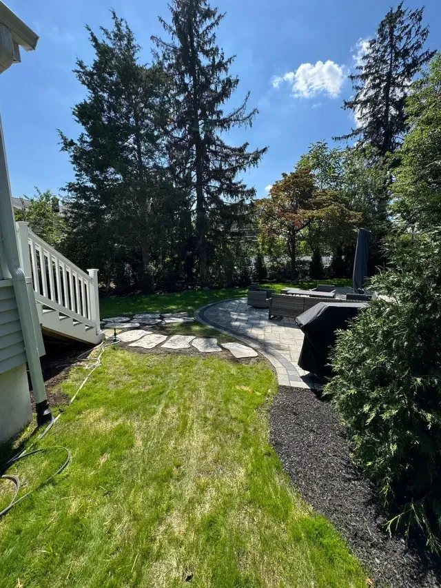 Sunny backyard with lawn, stone path, tall evergreens, and a house deck on the left