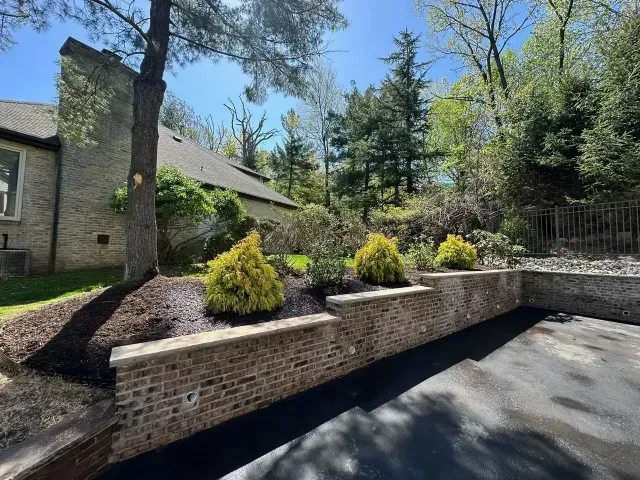 Brick retaining wall with yellow shrubs beside a stone house and driveway on a sunny day