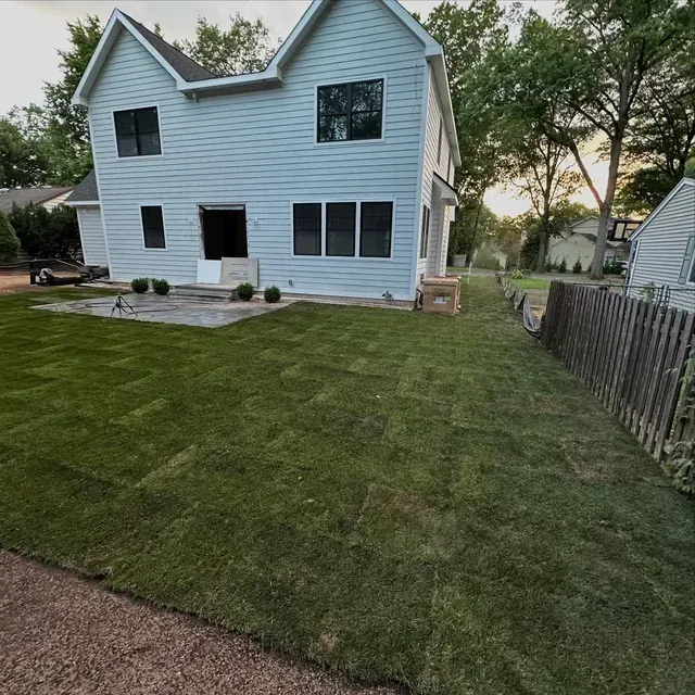 Backyard view of a light blue two-story house with a green lawn and wooden fence at dusk