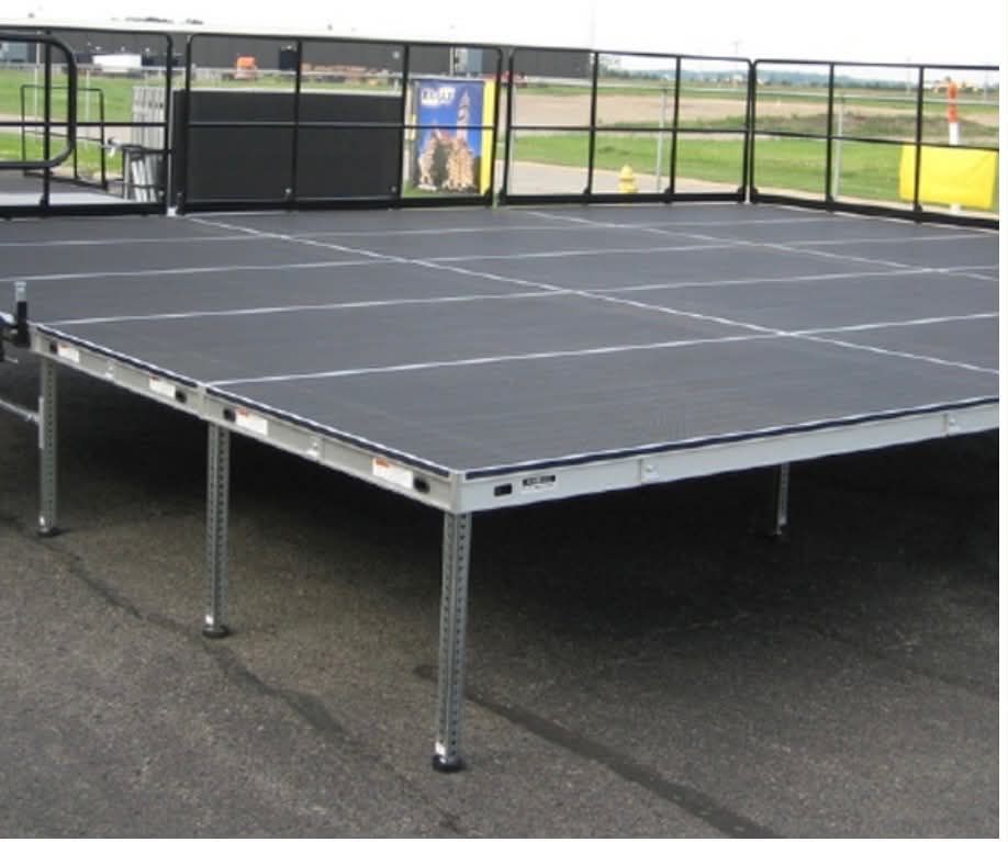 Raised black stage platform with metal legs on a paved surface, surrounded by fencing outdoors