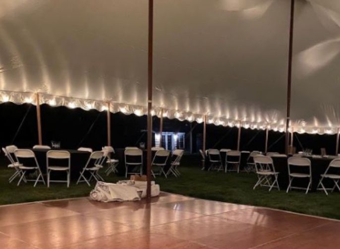 White tent with string lights, folding chairs, and tables on grass beside a wooden dance floor at night