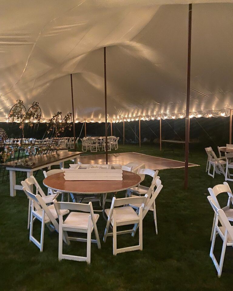Round tables and white folding chairs under a large tent lit with string lights at night.