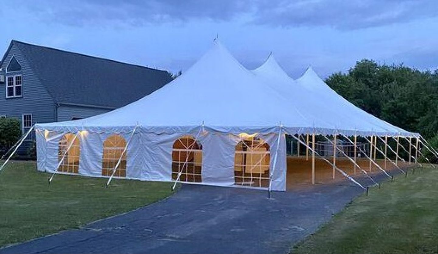 Large white event tent with peaked tops set up on a driveway beside a house at dusk.