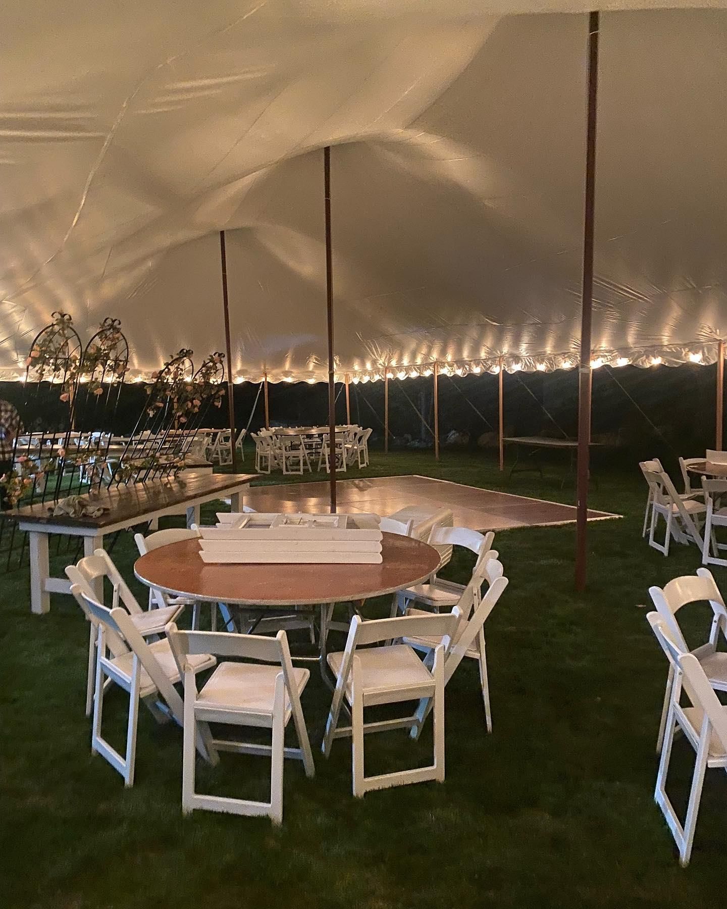 Round wooden tables and white chairs inside a lit tent at an outdoor evening event