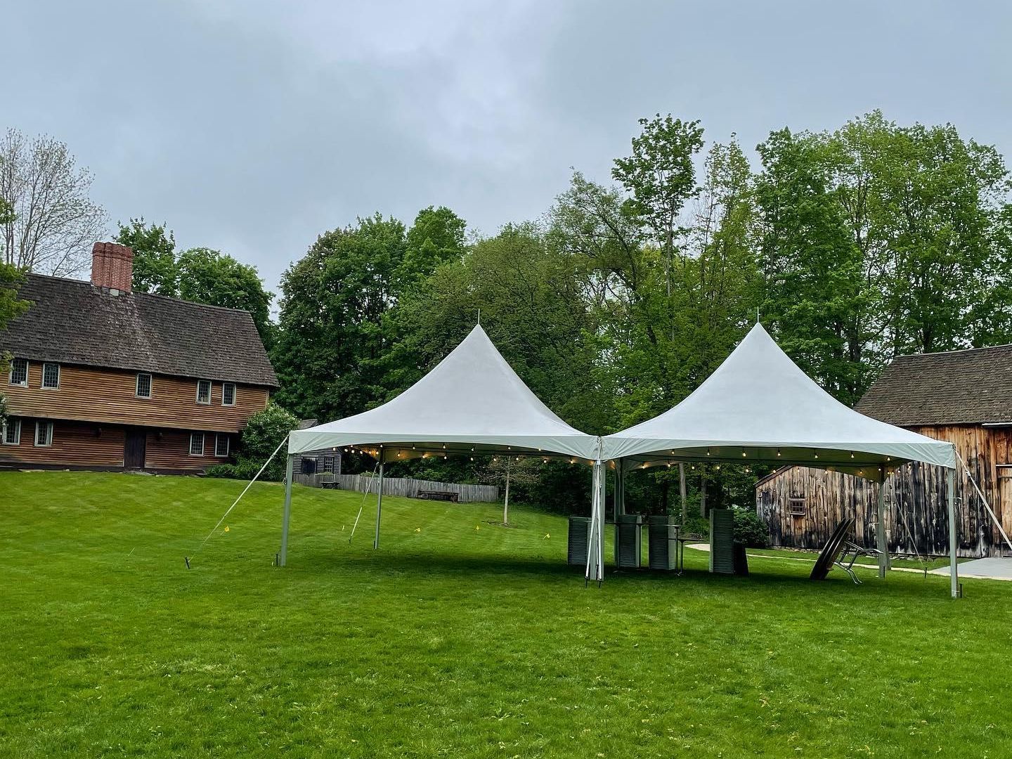 Two white canopy tents on a grassy lawn beside rustic buildings and trees