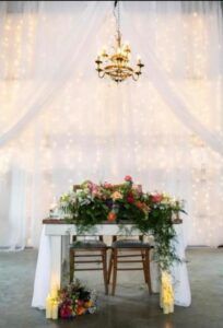Wedding arch with white drapes, chandelier, and floral-decorated table and chairs