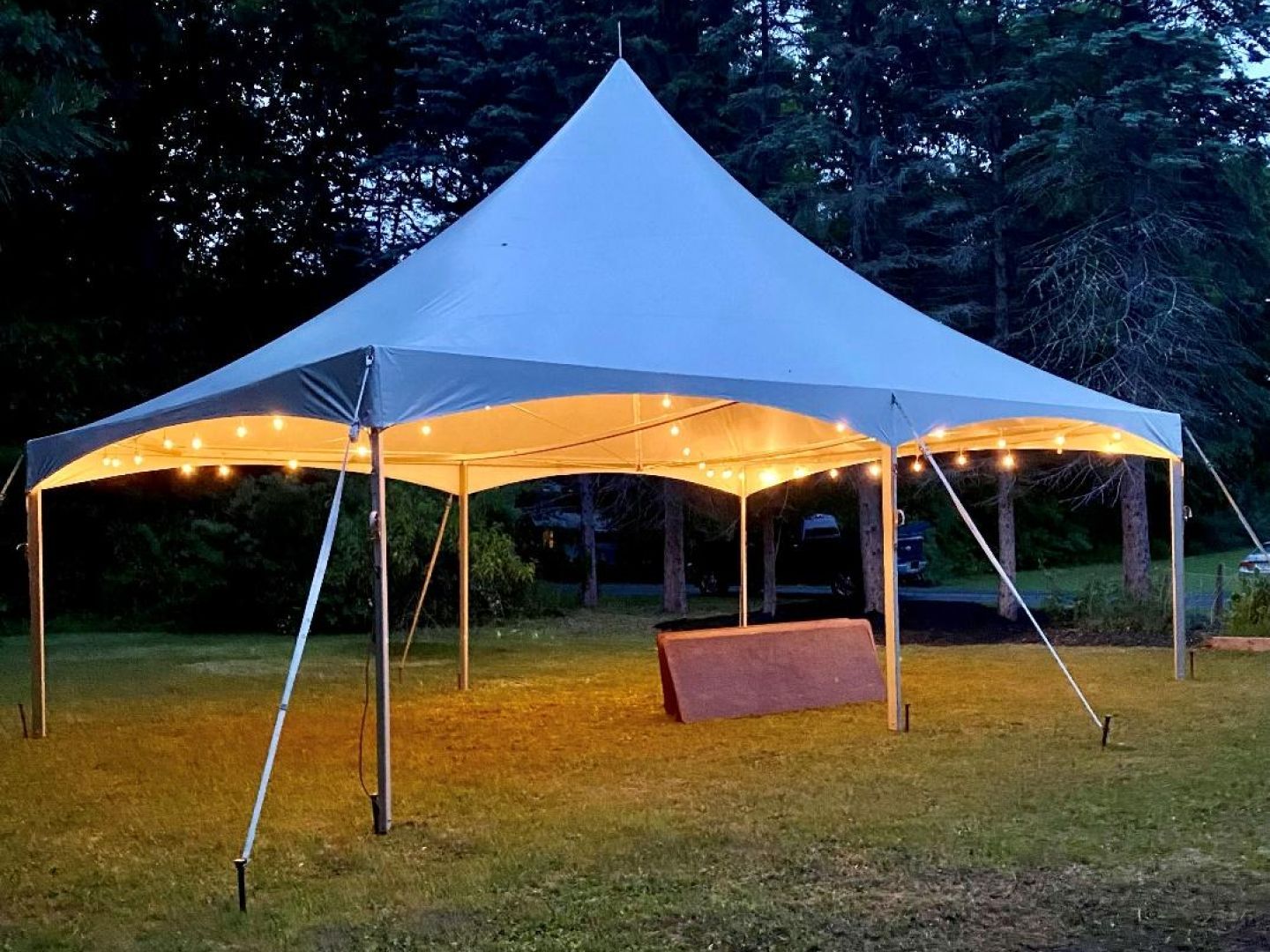 Lit white event tent on grass at dusk, glowing warmly with dark trees in the background.