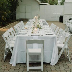 Long outdoor dining table with white chairs and linens in a backyard garden setting
