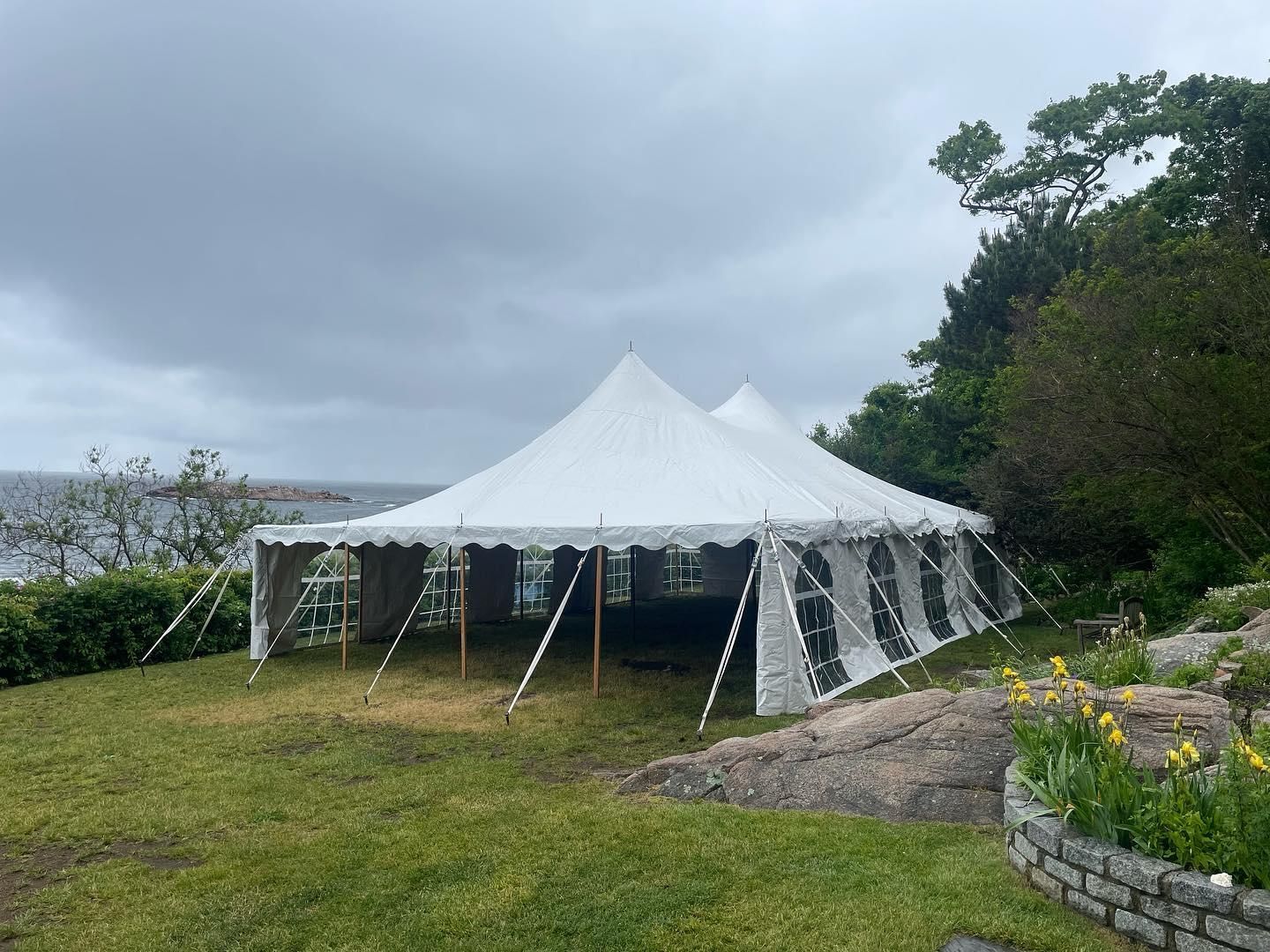 White event tent on a grassy lawn under a cloudy sky, with trees and a stone planter nearby