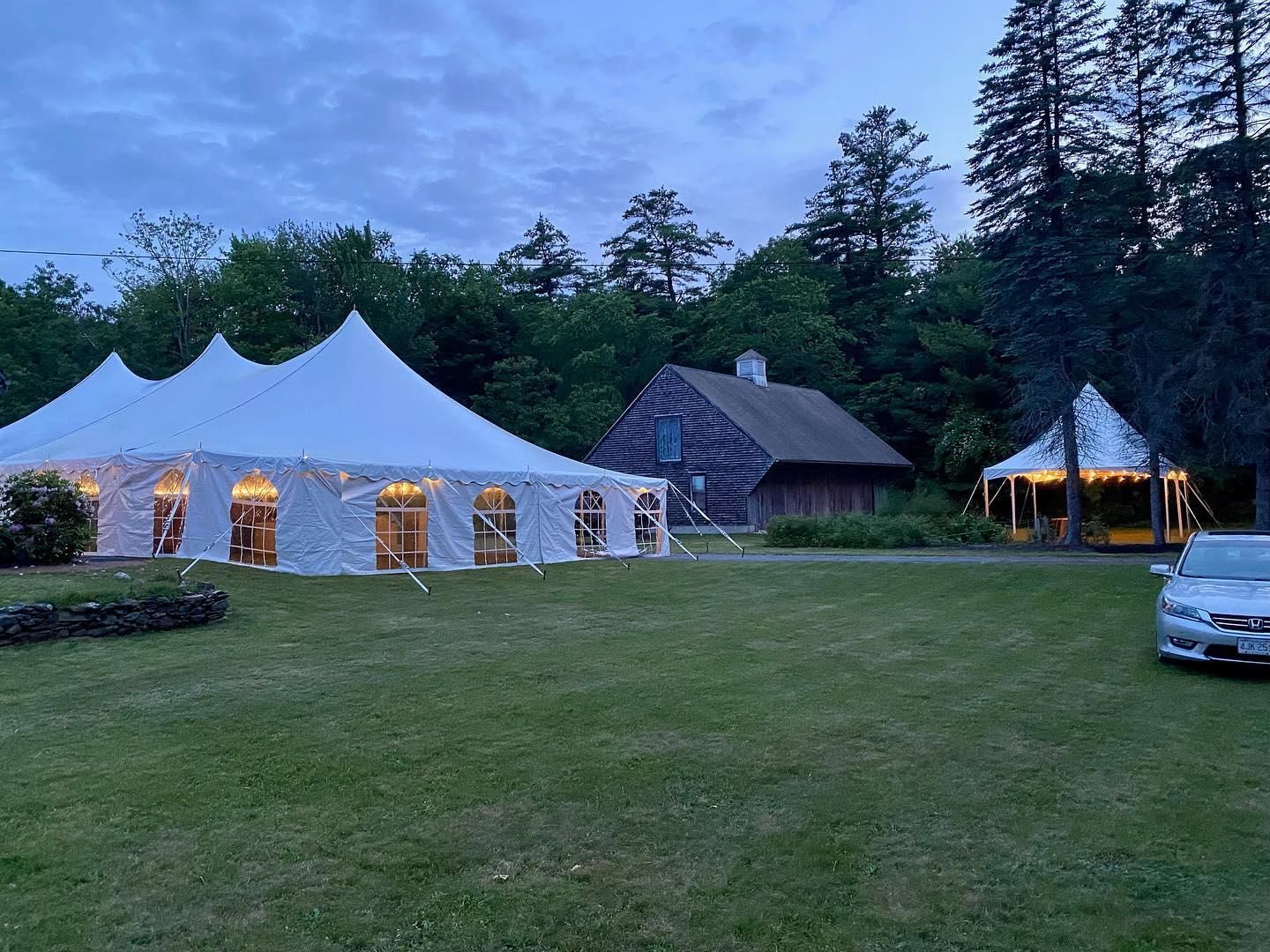 White event tent lit at dusk beside a rustic cabin and trees, with a grassy lawn and parked car.