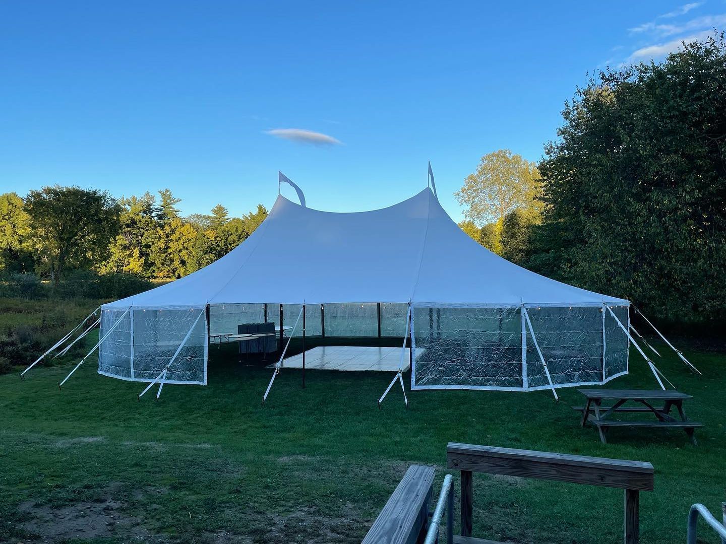 Large white event tent on a grassy field at sunset, with trees and picnic benches around it.