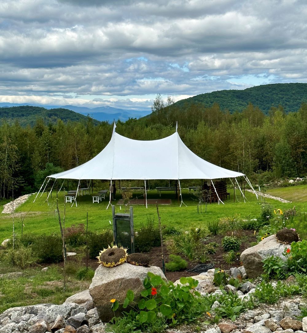 White event tent in a green mountain meadow under cloudy skies, with rocks and flowers in the foreground