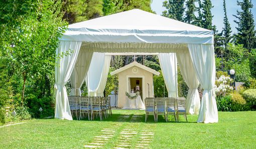 White wedding canopy with draped curtains and chairs on a grassy lawn, framed by trees and a small chapel backdrop