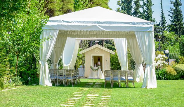 White wedding canopy with draped curtains and chairs on a grassy lawn, framed by trees and a small chapel backdrop