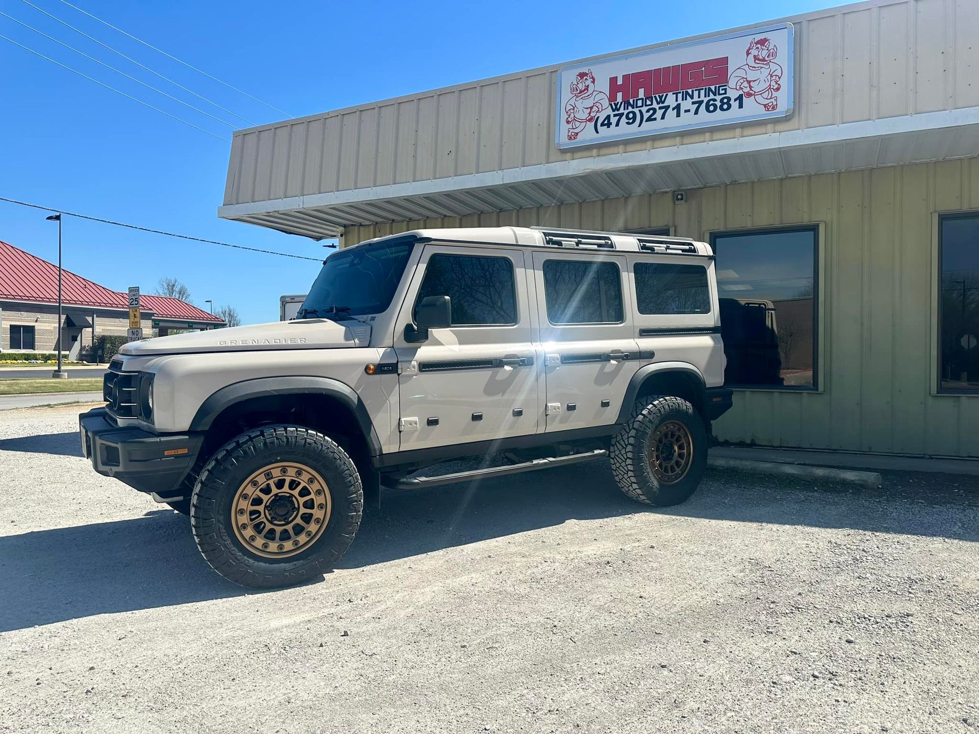 Tan Ineos Grenadier SUV parked in front of a building with a sign.