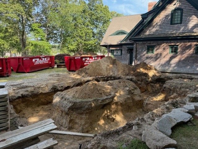 Excavation site next to a brown house with large piles of dirt and red dumpsters.