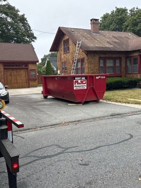Red dumpster in a driveway in front of a house, a ladder propped on the roof.