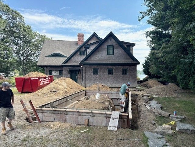 Construction site with two men. A house with cedar shingles is in the background. A red dumpster and sand piles are present.