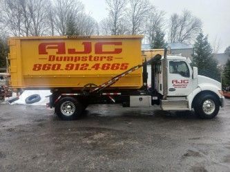 Metal dumpster filled with junk, set on a brick patio with green bins in the background.