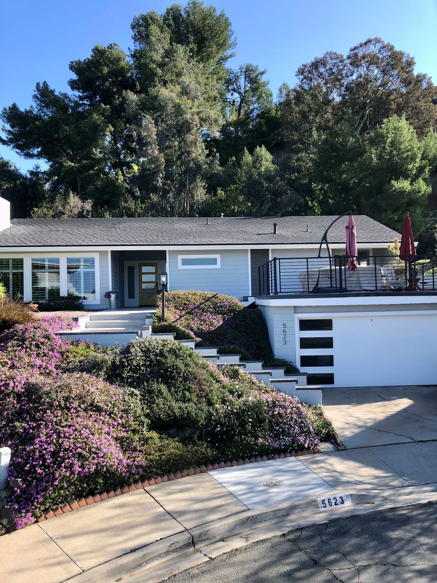 House with blue siding, gray roof, and white garage door, surrounded by purple flowering bushes.