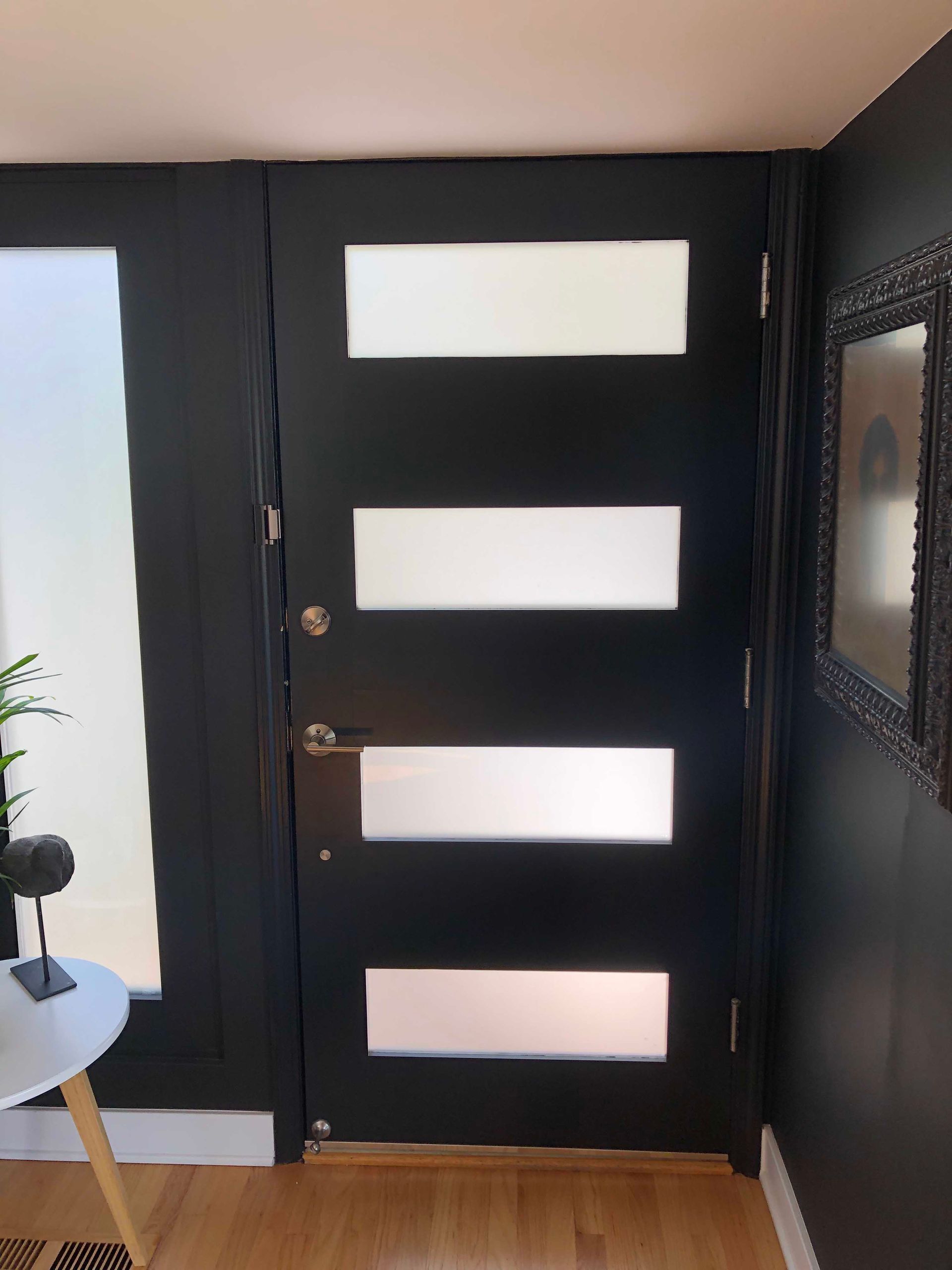 Black front door with four frosted glass panels, next to a window and wall with a framed art piece.