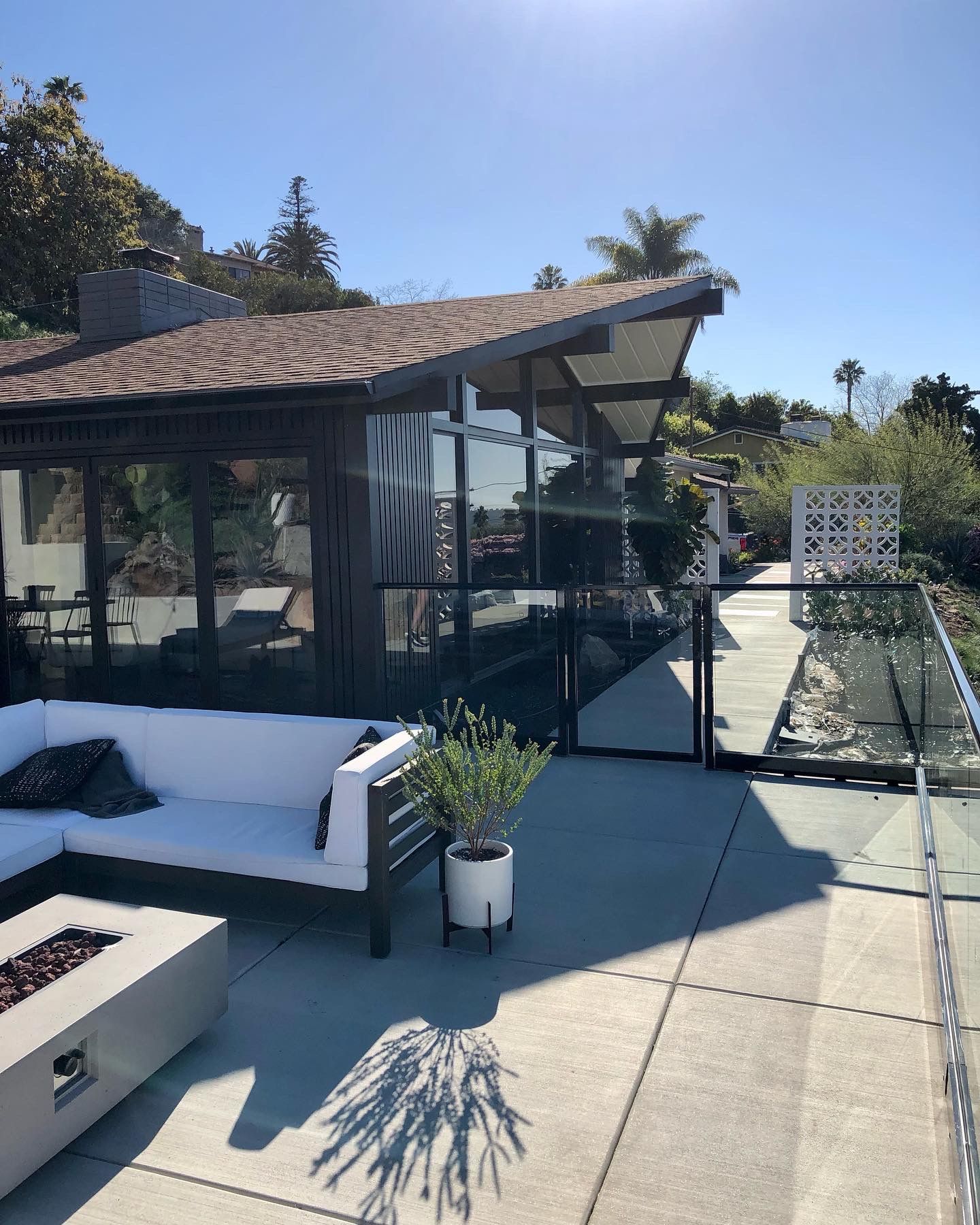 Modern home with outdoor patio. White sofa, fireplace, potted plant. Glass wall, tiled roof, sunny day.