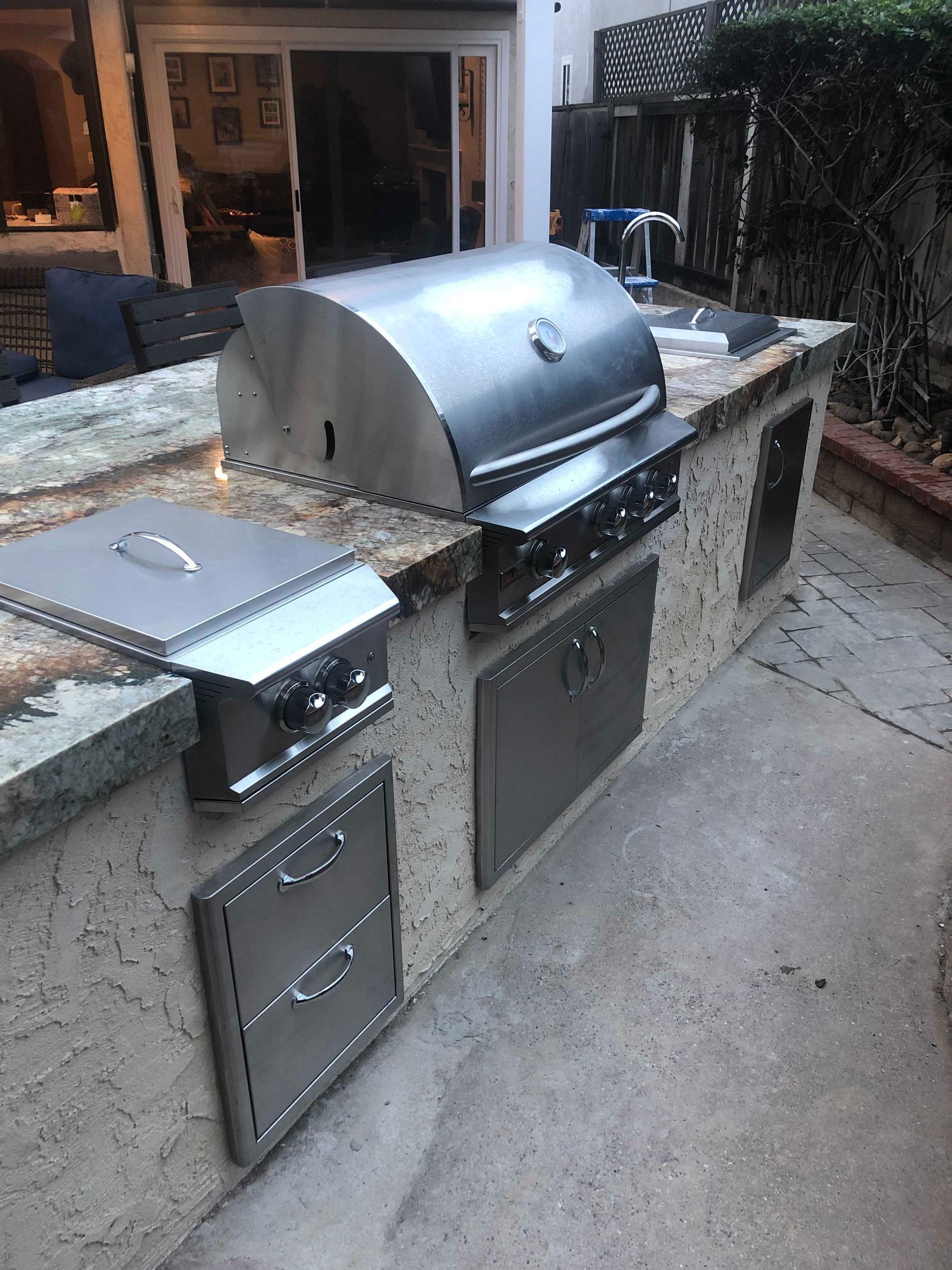 Outdoor built-in kitchen with stainless steel grill and side burner, granite countertop, concrete base, and storage doors.