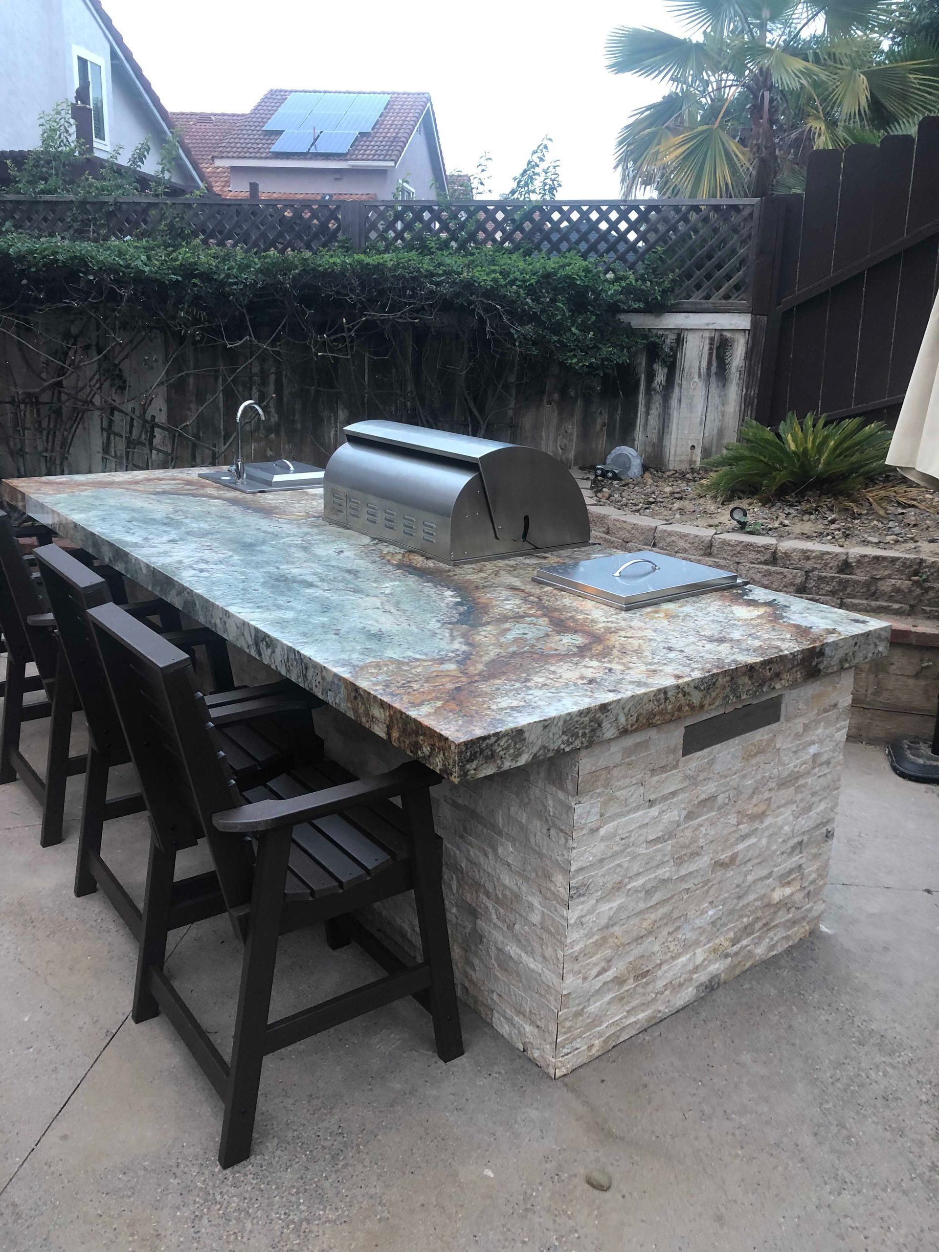 Outdoor kitchen island with grill, seating, and stone facade in a backyard setting.