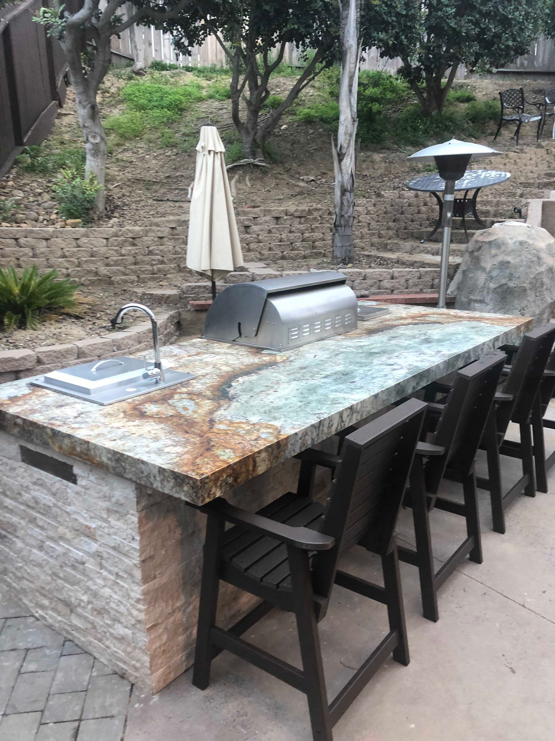 Outdoor kitchen island with a granite countertop, grill, sink, and bar seating.