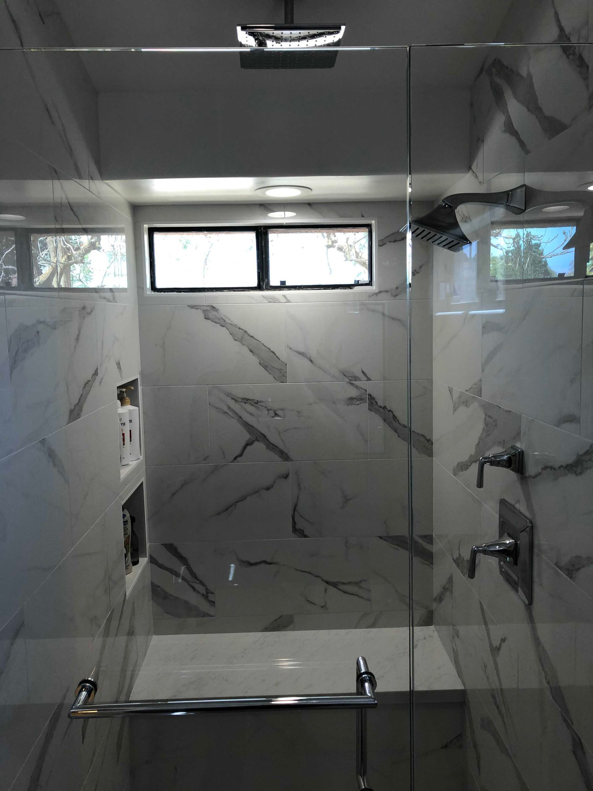 Modern bathroom shower with marble-like tile, glass door, window, and chrome fixtures.