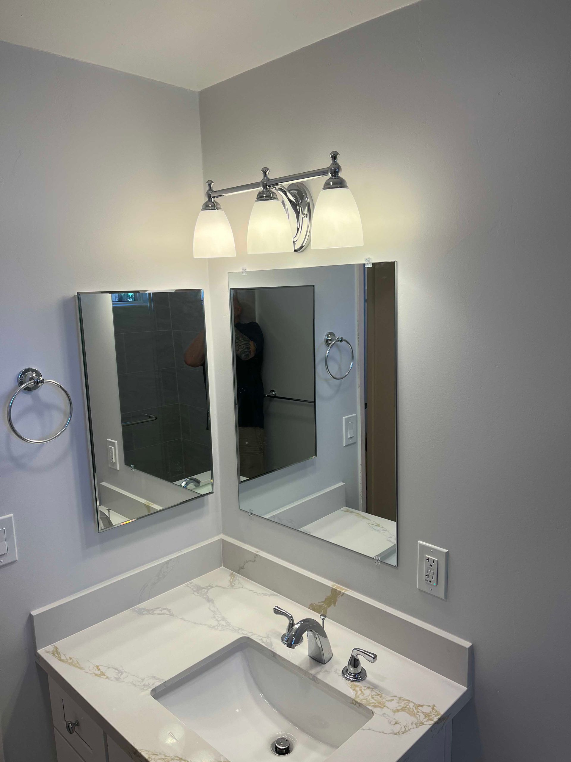 Bathroom with three-bulb light fixture above mirrors and a sink. The walls are gray, and the countertop is white.