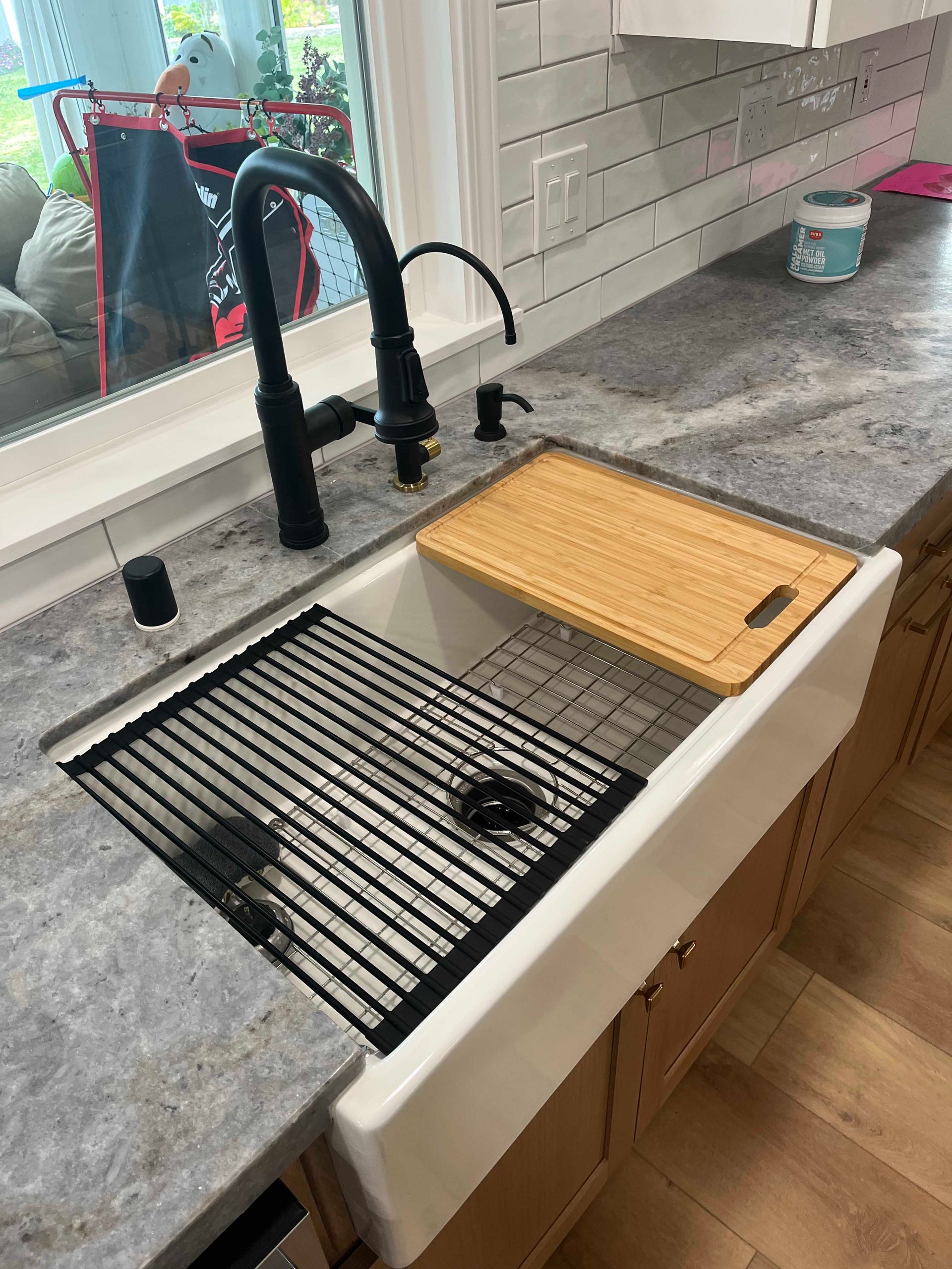 Black faucet over a farmhouse kitchen sink with a cutting board and a dish rack on gray countertops.