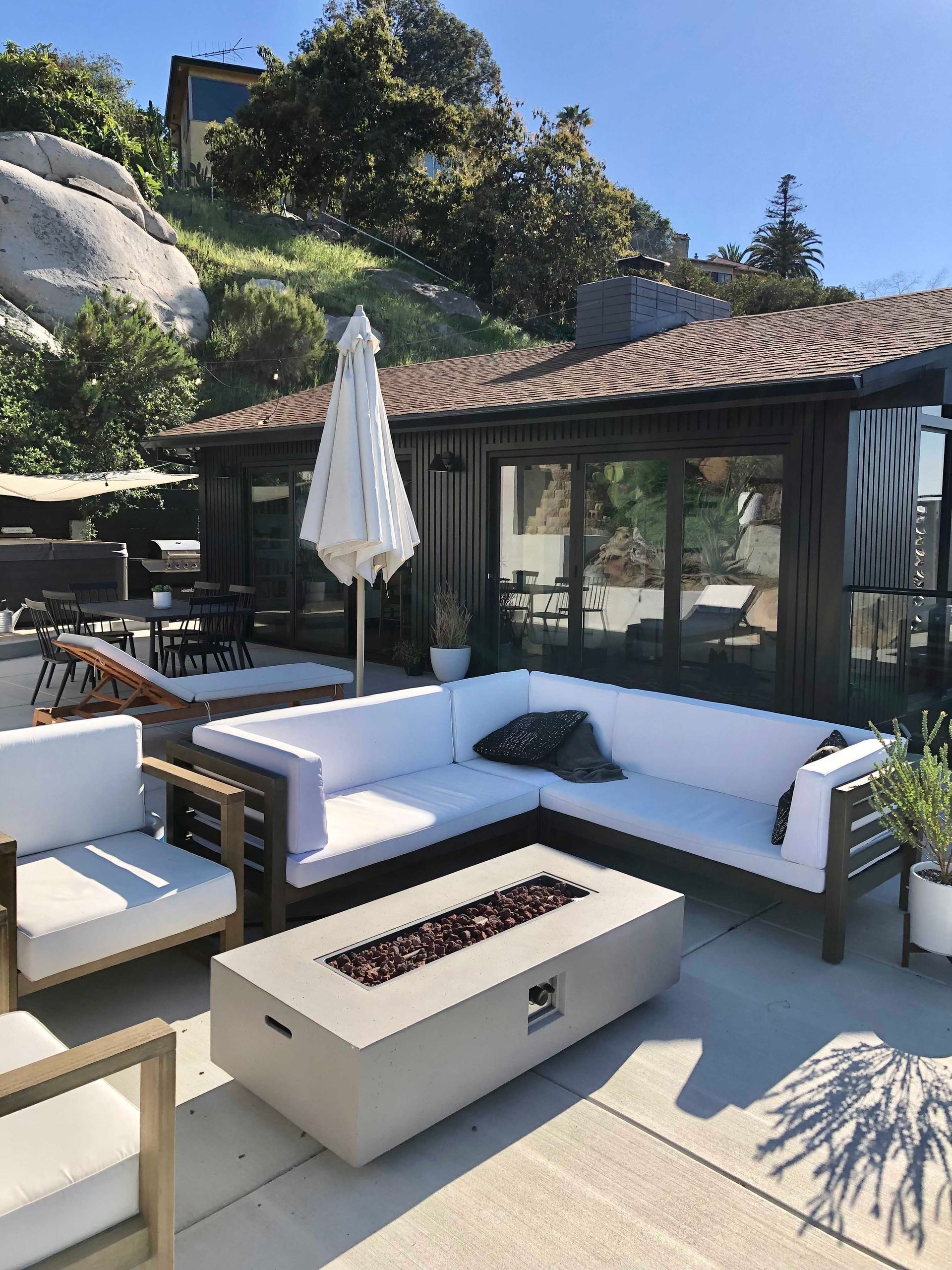 Outdoor patio with white sectional sofa, fire pit, and umbrella in front of a house, rocks and trees in background.